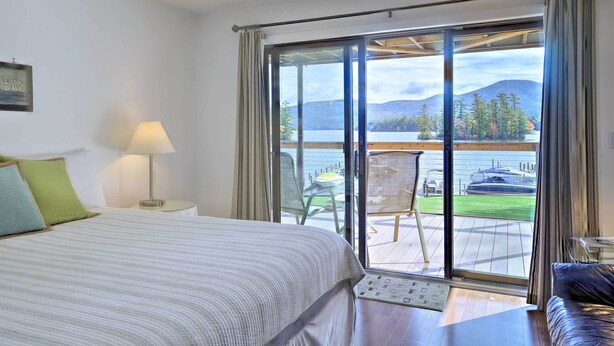 A hotel room with a bed, a nightstand, a lamp, and a view of a lake and mountains through a sliding glass door. - Bolton Landing Suite w/ Deck, Lake George Access (Bolton Landing)