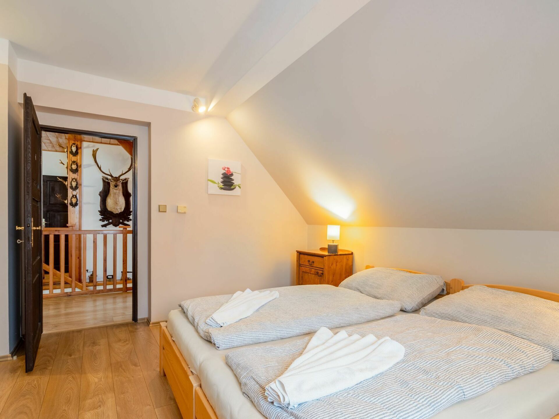 A bedroom with two beds, a wooden headboard, a nightstand, a lamp, and a deer head mounted on the wall.