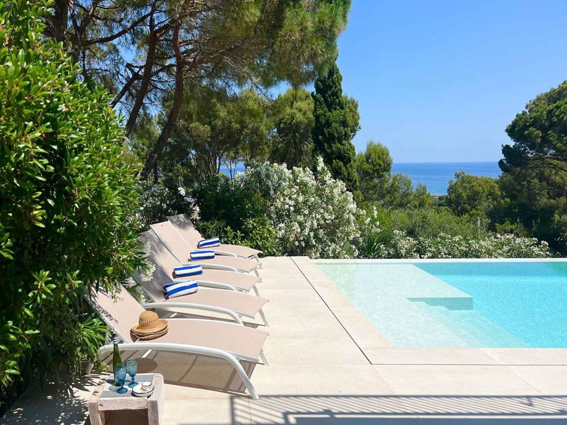 A poolside area with lounge chairs and a table, surrounded by greenery and overlooking a body of water.