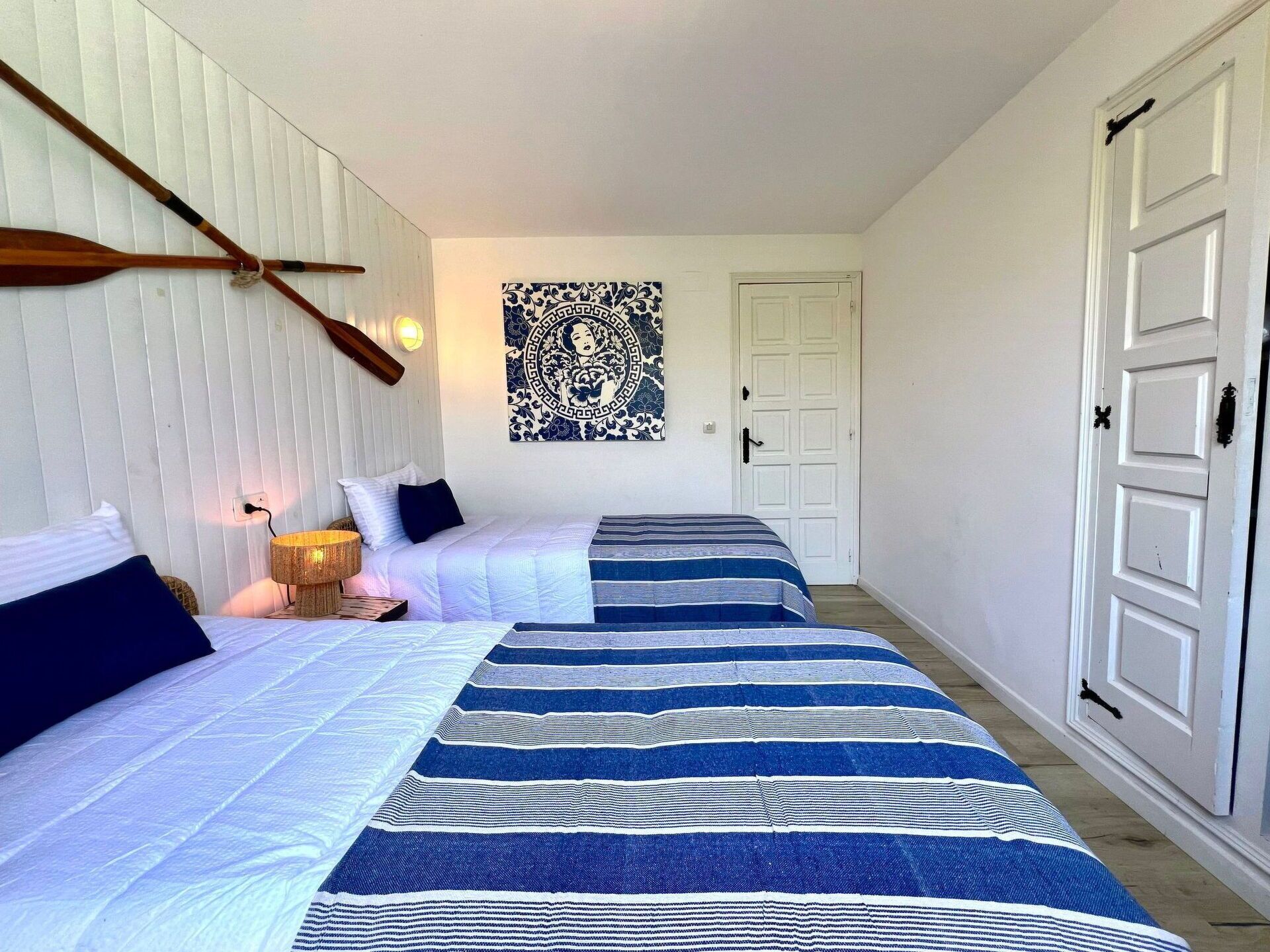 Une chambre avec deux lits, une pagaie en bois fixée au mur, une tapisserie décorative et une porte blanche.