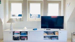 Un salon moderne avec un téléviseur à écran plat posé sur un meuble blanc, des étagères, une lampe et une vue sur la ville grâce à de grandes fenêtres.