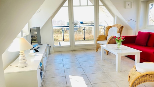Une pièce moderne et bien éclairée, avec un canapé rouge, des fauteuils en osier, une table blanche et une grande fenêtre donnant sur l’extérieur.