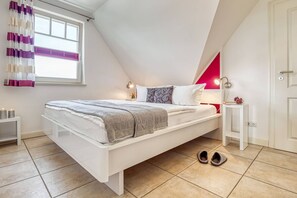 A modern bedroom with a sloped ceiling, a bed with white bedding, a bedside table with a lamp, and a window with curtains. - Comfortable Boddenstern vacation apartment in Zingst with balcony and WLAN - (Zingst)