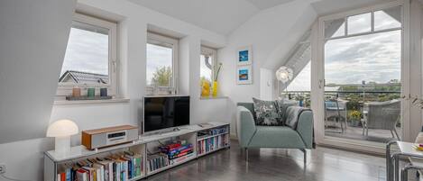 A modern living room with a flat-screen TV, bookshelves, a sofa, and a balcony with outdoor furniture.