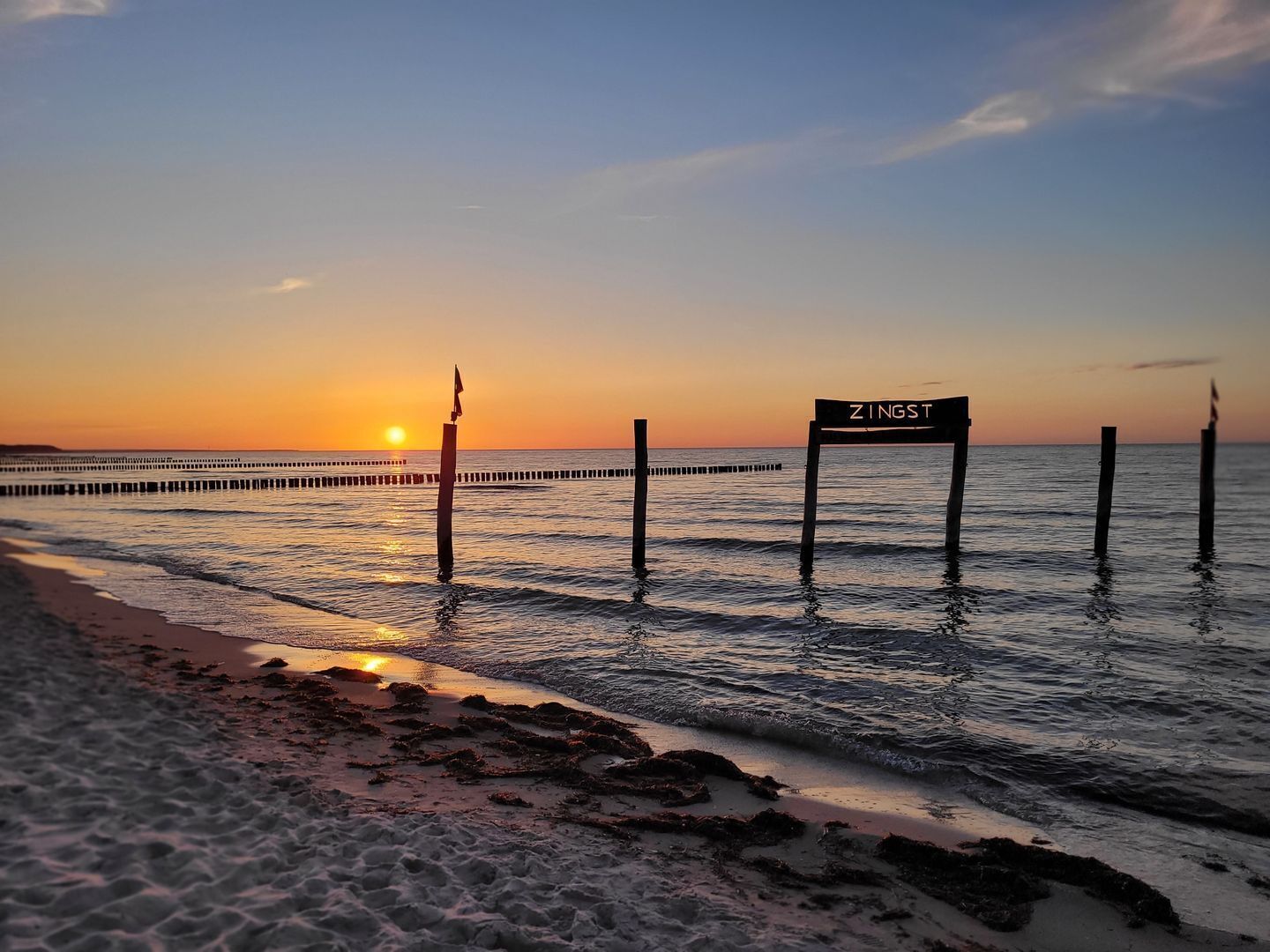 Escena de platja amb un moll que s'endinsa a l'aigua, un cartell que diu 'ZINGST' i el sol ponent-se al fons.