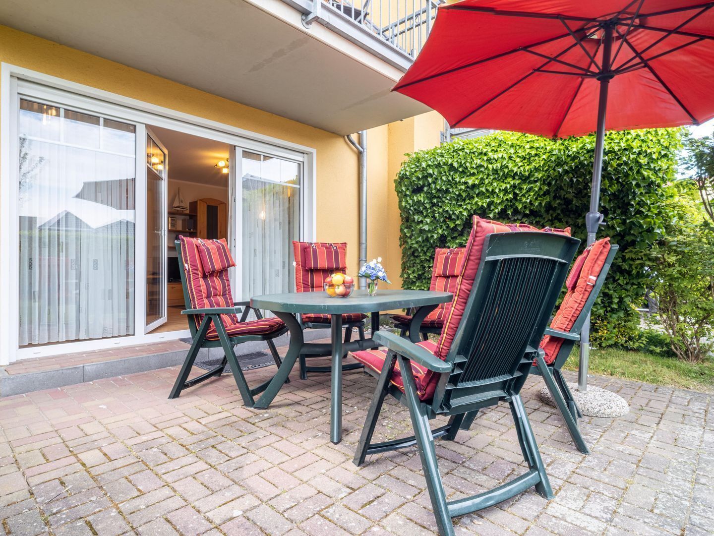 An outdoor patio with a table and chairs, a red umbrella, and a hedge in the background.