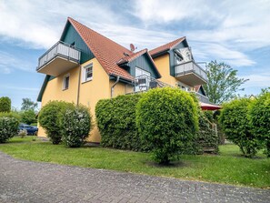 Een huis met twee verdiepingen, een rode dakbedekking, een balkon op de bovenverdieping en een verzorgde tuin met hagen.