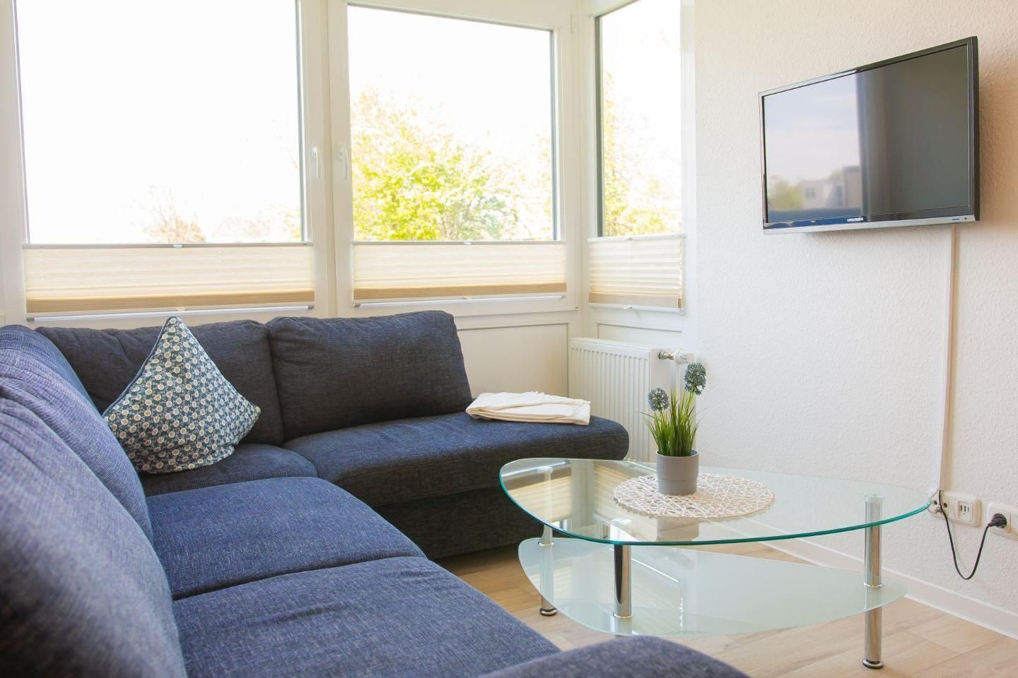 Een woonkamer met een bank, een glazen salontafel, een flatscreen televisie aan de muur en een raam met jaloezieën.