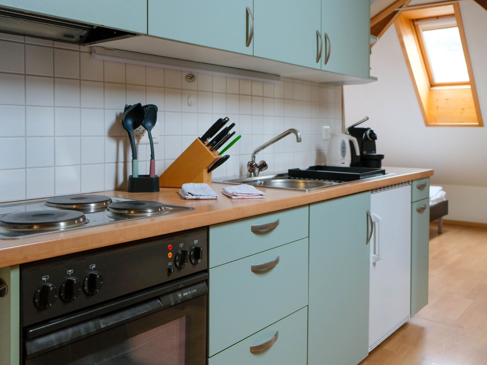 Een keuken met een fornuis, oven, spoelbak en lichtgroene kastjes. Op het aanrecht staan keukengerei en een messenblok.