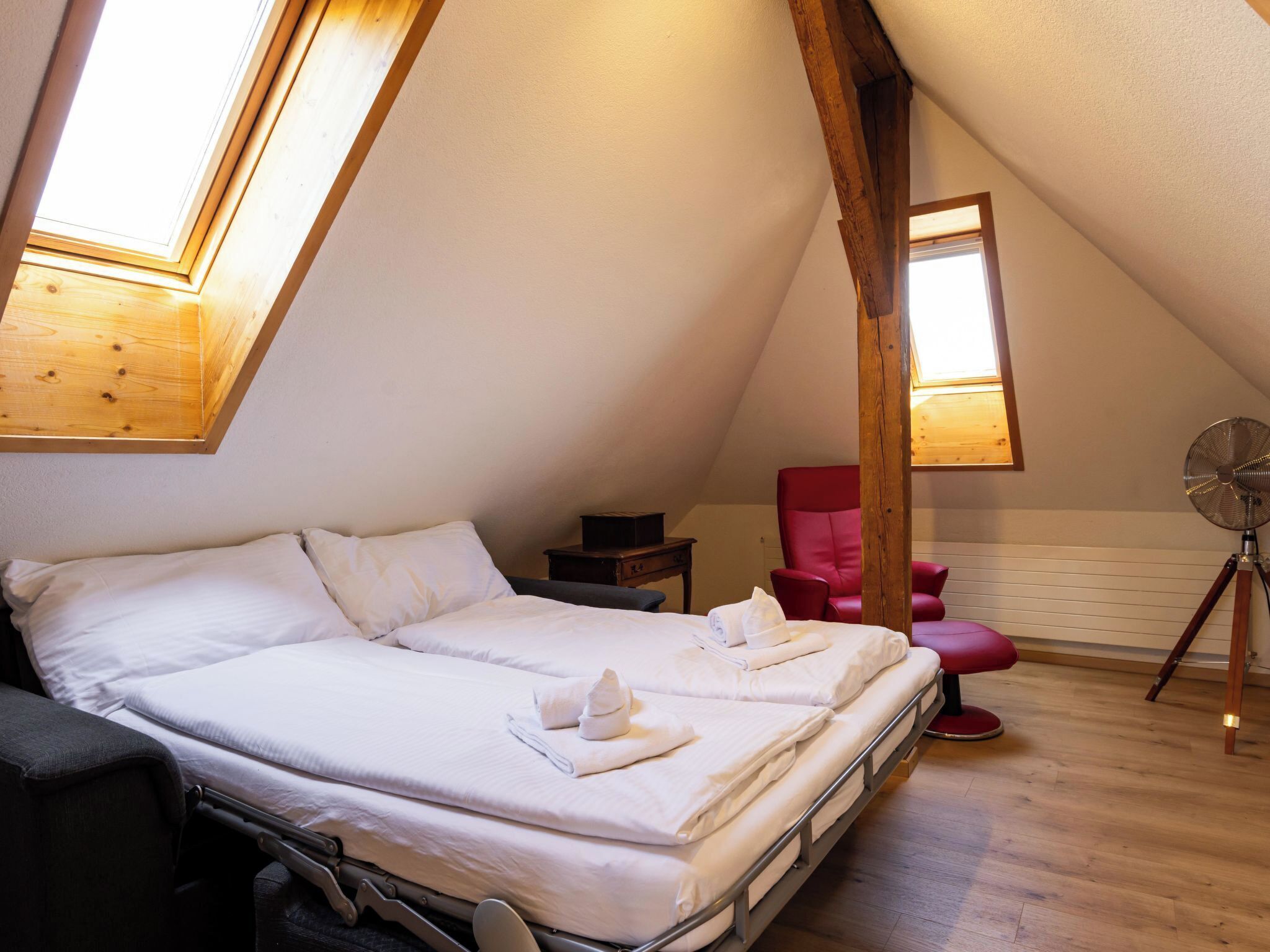 A bedroom with a bed, a red chair, a fan, and a wooden ceiling structure.