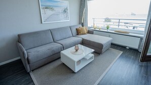 Een moderne woonkamer met een grijze modulaire bank, een witte salontafel en een groot raam met uitzicht op zee.