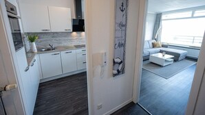 Een moderne keuken met witte kastjes en een grijze achterwand, een zithoek met een grijze bank en een salontafel, en een groot raam dat zorgt voor natuurlijk licht.