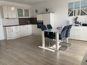 A modern kitchen with white cabinets, a wooden backsplash, and a dining area with a glass table and black chairs. - Lee - Am Kampland - Herzlich Willkommen in Dahme! (Dahme)