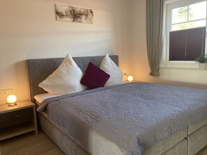 Una cama bien hecha con un edredón gris, dos almohadas blancas y una almohada roja. Mesitas de noche con lámparas a cada lado de la cama. Un cuadro cuelga en la pared sobre la cama.