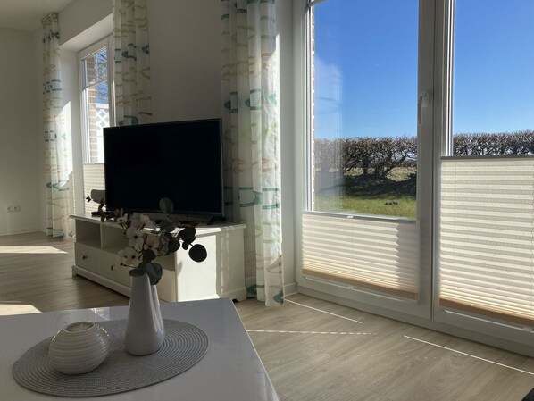 Un salon moderne avec un téléviseur à écran plat posé sur un meuble blanc, un vase de fleurs sur une table et de grandes fenêtres avec des rideaux.