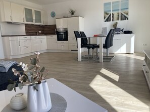 A modern kitchen with white cabinets, a built-in oven, and a dining area with a table and chairs. - Lee - Am Kampland - Herzlich Willkommen in Dahme! (Dahme)