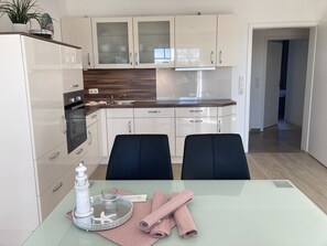 Cocina moderna con armarios blancos, salpicadero de madera y mesa de comedor de vidrio, con un adorno blanco y toallas rosas.