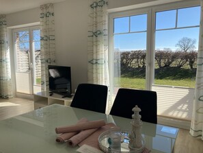 Una habitación con una mesa de vidrio, dos sillas, un televisor y vistas a un jardín a través de amplias ventanas.