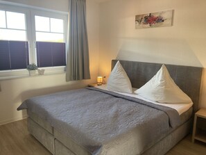 Una cama bien hecha con cabecera gris y almohadas, una lámpara de noche y una ventana con persianas y cortina.