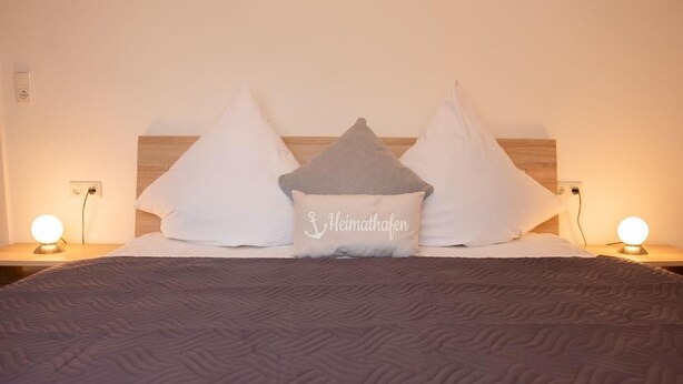 A neatly made bed with a textured blanket, two bedside lamps, and a pillow with the text "Heimathafen". - Kleine Perle - Waldstraße - "Relaxation and recreation in the little pearl on (Dahme)