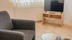A living room with a grey sofa, a glass coffee table with decorative items, and a TV on a wooden stand. - Kleine Perle - Waldstraße - "Relaxation and recreation in the little pearl on (Dahme)