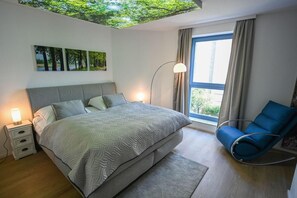 Un dormitorio con cama, lámparas de noche, un sillón azul, una lámpara de pie y una ventana con cortinas.