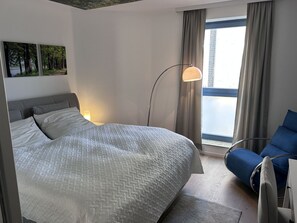 Ein Hotelzimmer mit einem großen Bett, einem Schreibtisch, einem Sessel, einer Stehlampe und einem Fenster mit Vorhängen.