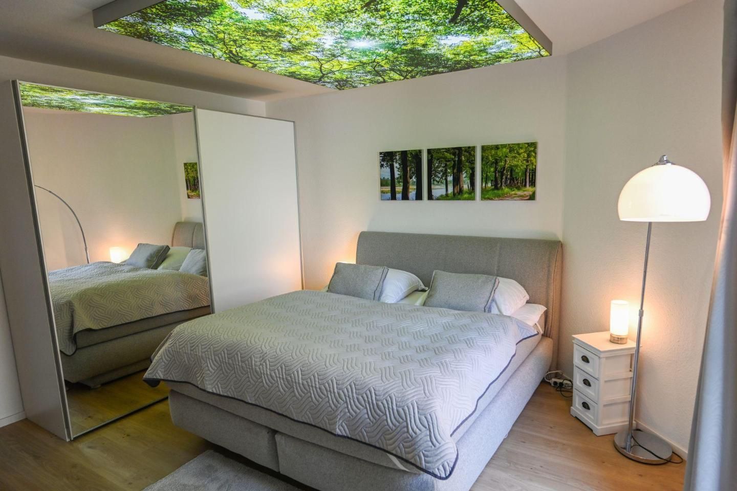 A modern bedroom with a large bed, a bedside table, a floor lamp, and a ceiling with a tree design.