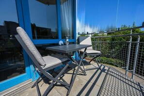 Ein Balkon mit Tisch und zwei Stühlen, ein Fenster mit Vorhängen und Blick auf Grünflächen.