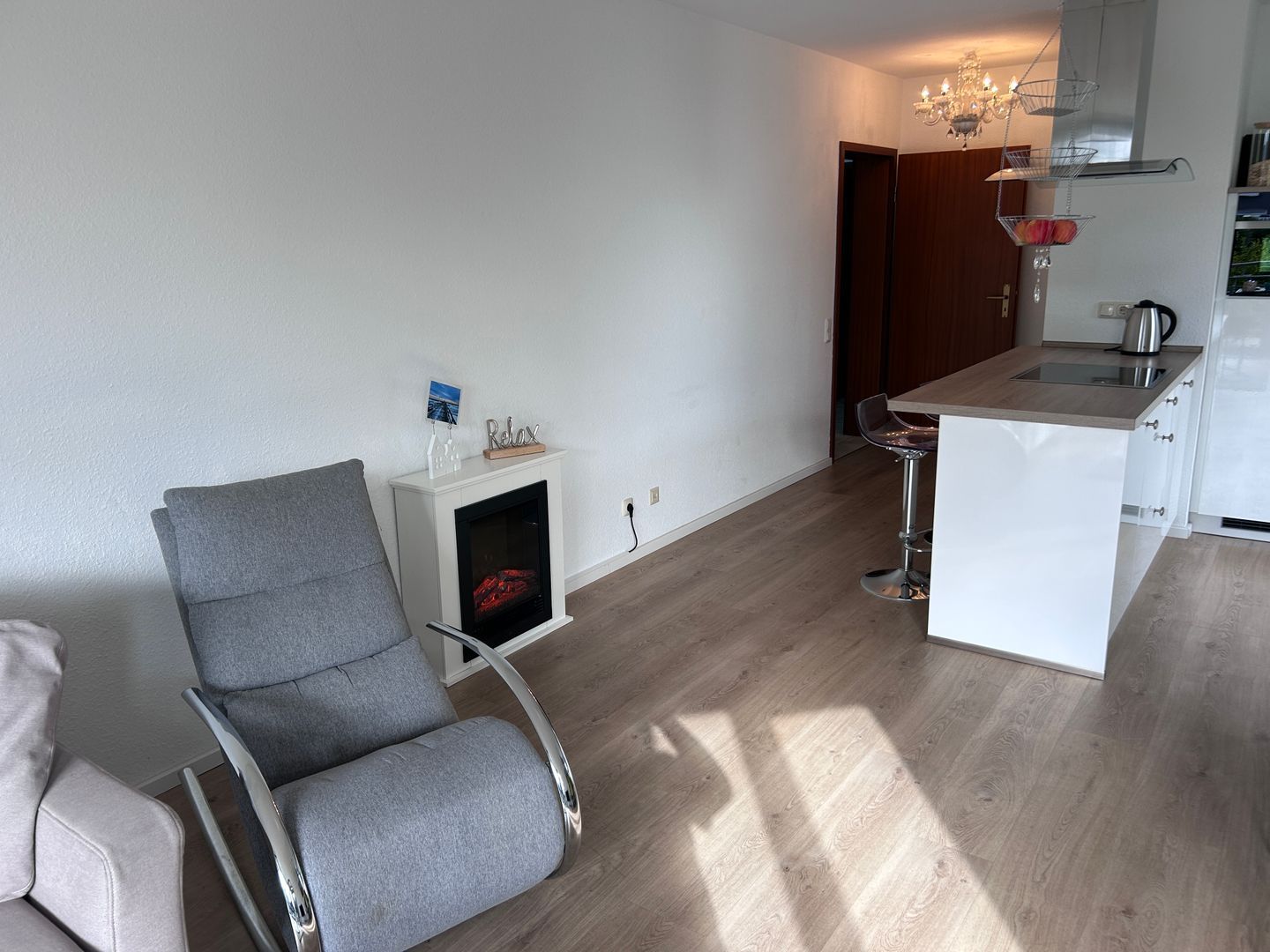 Un salon moderne avec un fauteuil gris, une petite cheminée blanche et un coin cuisine comprenant un réfrigérateur blanc et une bouilloire.