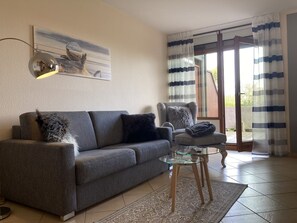 A living room with a grey sofa, a glass coffee table, a fluffy grey pillow, a painting on the wall, and a view through a glass door with striped curtains. - Lindenhof - Gentle wave - "Gentle wave"... Beach chair on the beach included! (Dahme)