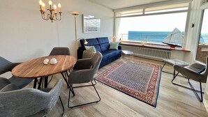 A modern living room with a round wooden table, grey upholstered chairs, a blue sofa, and a patterned rug. A large window offers a view of the sea. - Beach hotel - 56 - sea view and beach chair included! (Dahme)