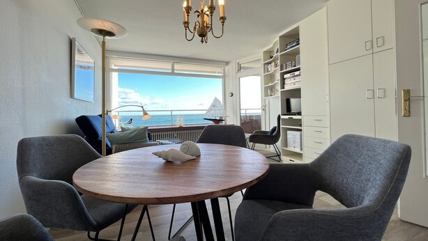 A modern dining area with a round wooden table, grey upholstered chairs, a chandelier, and a view of the sea through large windows. - Beach hotel - 56 - sea view and beach chair included! (Dahme)