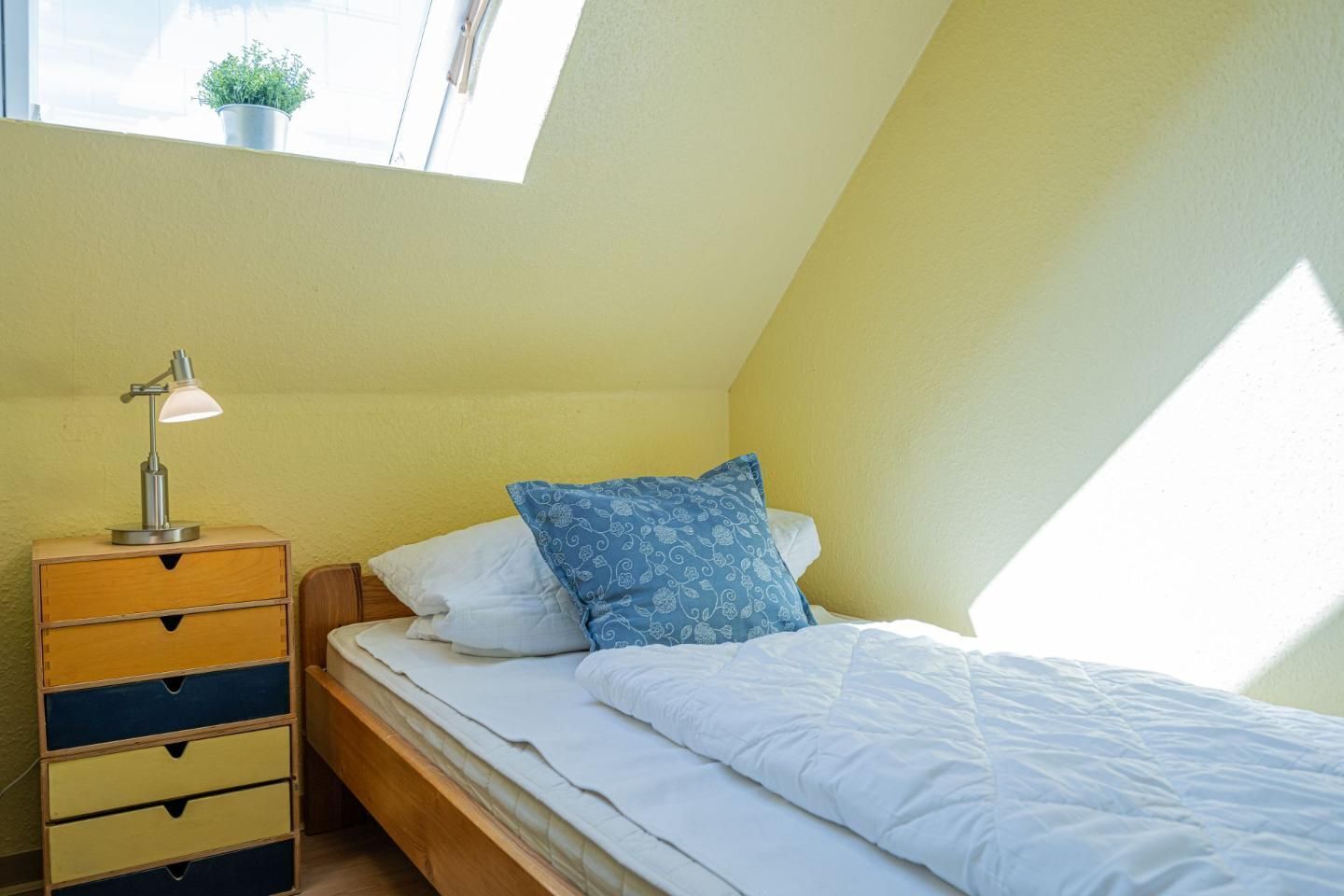 Een knus slaapkamer met een enkel bed, een nachtkastje met lamp, en een raam met uitzicht.