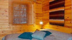 Une chambre dans un chalet en bois, avec un lit, une table de chevet, une lampe et une fenêtre avec des stores.