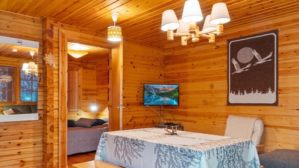 L’intérieur d’un chalet accueillant, avec des murs en bois, un téléviseur fixé au mur, une table à manger recouverte d’une nappe à motifs floraux, un banc et un canapé.