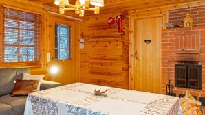 Une pièce chaleureuse aménagée dans un chalet en bois, avec un plafond en bois, une cheminée, une table à manger recouverte d’une nappe et un canapé agrémenté de coussins.