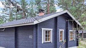 Une maison en bois sombre, portant le numéro 152 sur le côté, avec deux fenêtres dotées de volets blancs et une cheminée.