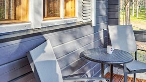 Une terrasse en bois avec une table ronde et deux chaises, une fenêtre avec des stores et une vue sur les arbres.