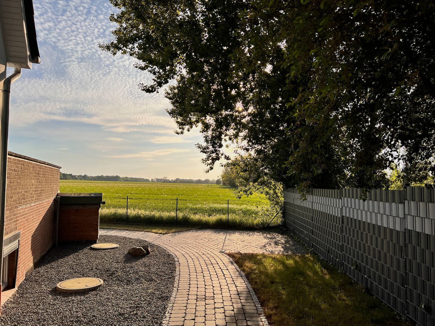 Ein gepflasterter Weg führt zu einem grünen Feld, einem Backsteinhaus und einem Metallzaun.