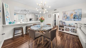 Eine moderne Küche mit weißen Schränken, eine Essecke mit Glastisch und Stühlen sowie ein Wohnzimmer mit Sofa und einer dekorativen Wandarbeit.