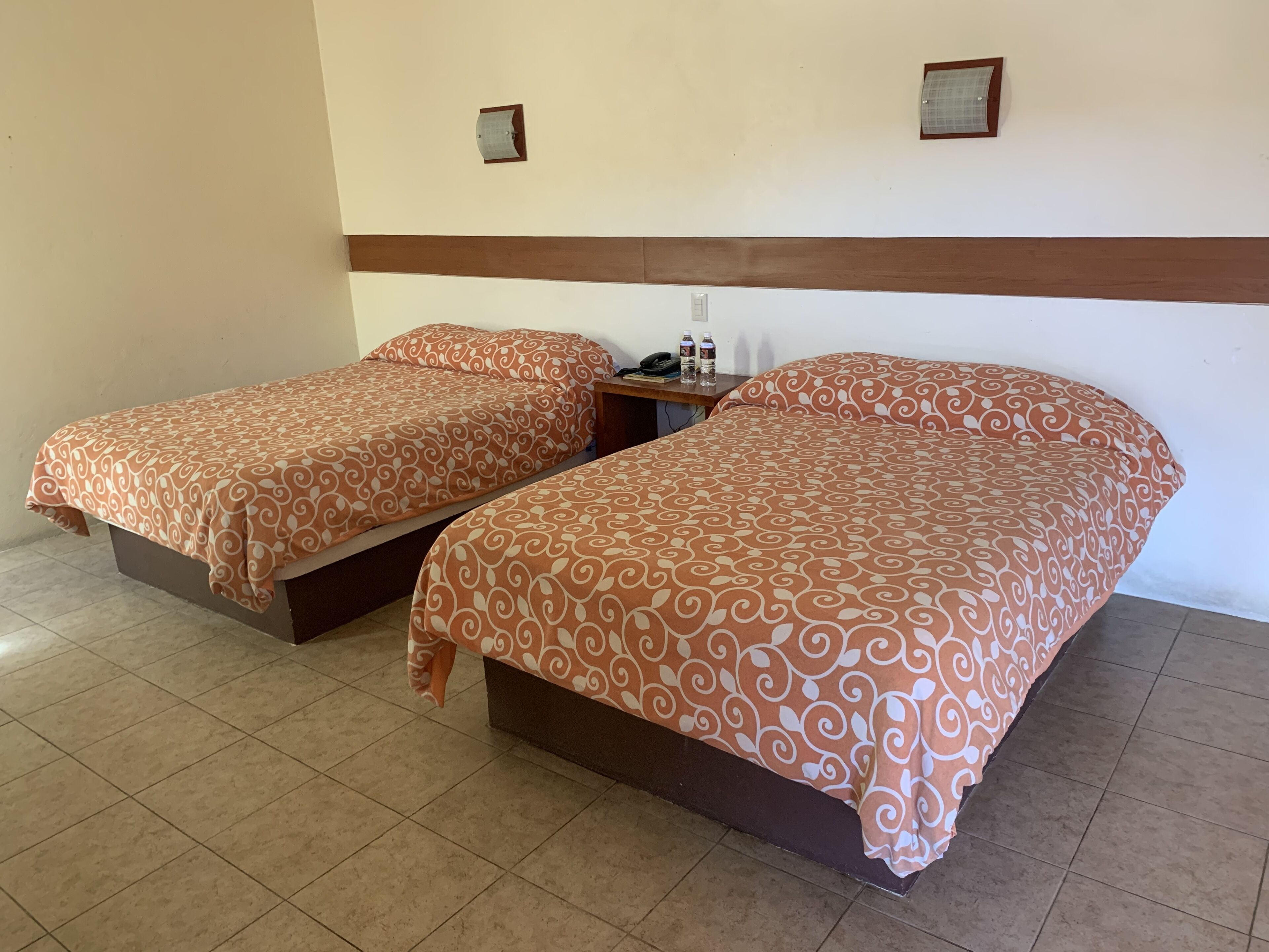 Une chambre d’hôtel avec deux lits, chacun recouvert d’une couvre-lit à motifs, une table de chevet avec un téléphone et une bouteille d’eau, et un luminaire fixé au mur.