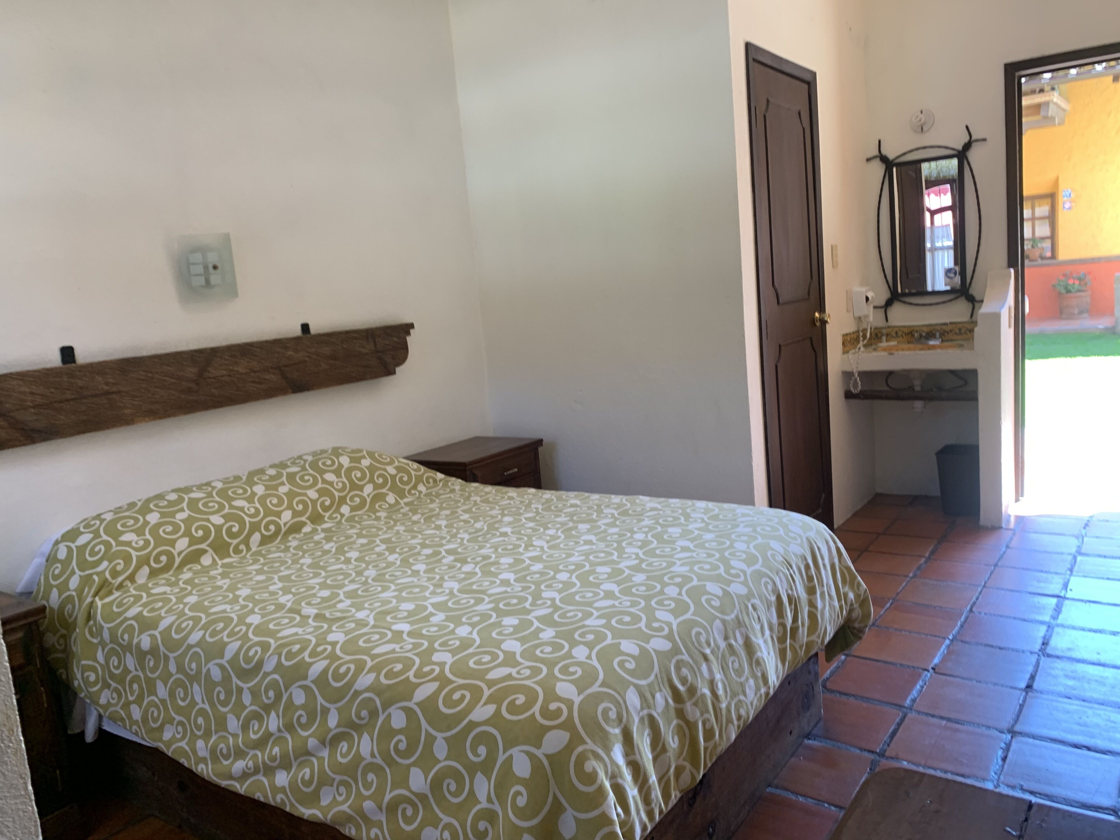 Un dormitorio con cama, cabecero de madera, mesita de noche pequeña, puerta y baño con lavabo y espejo.