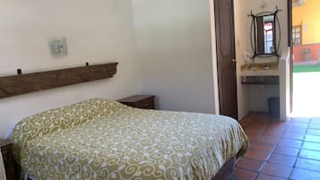 Un dormitorio con cama, cabecero de madera, mesita de noche pequeña, puerta y baño con lavabo y espejo.