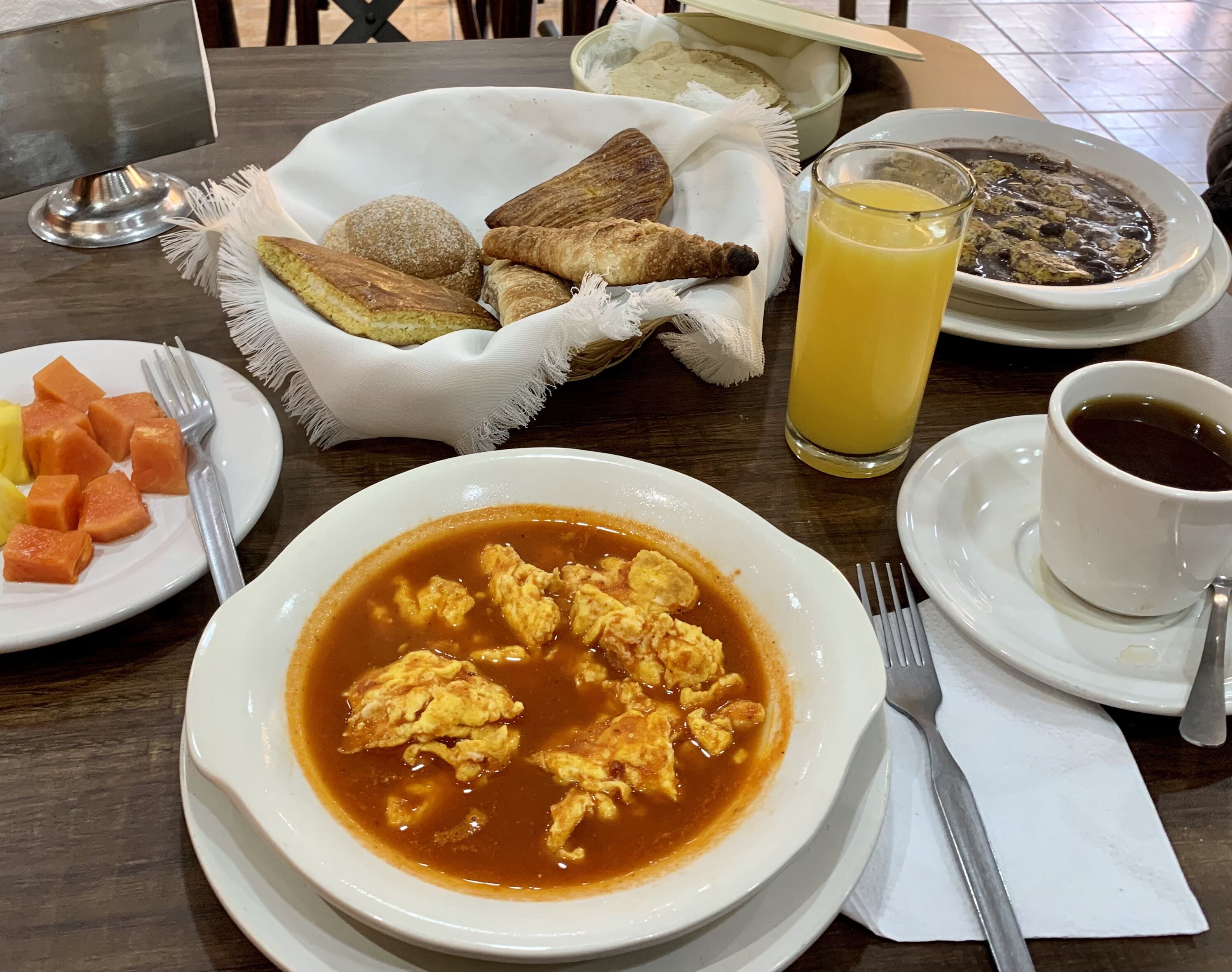 Una mesa con desayuno que incluye huevos revueltos, tostadas, fruta y un vaso de jugo de naranja.