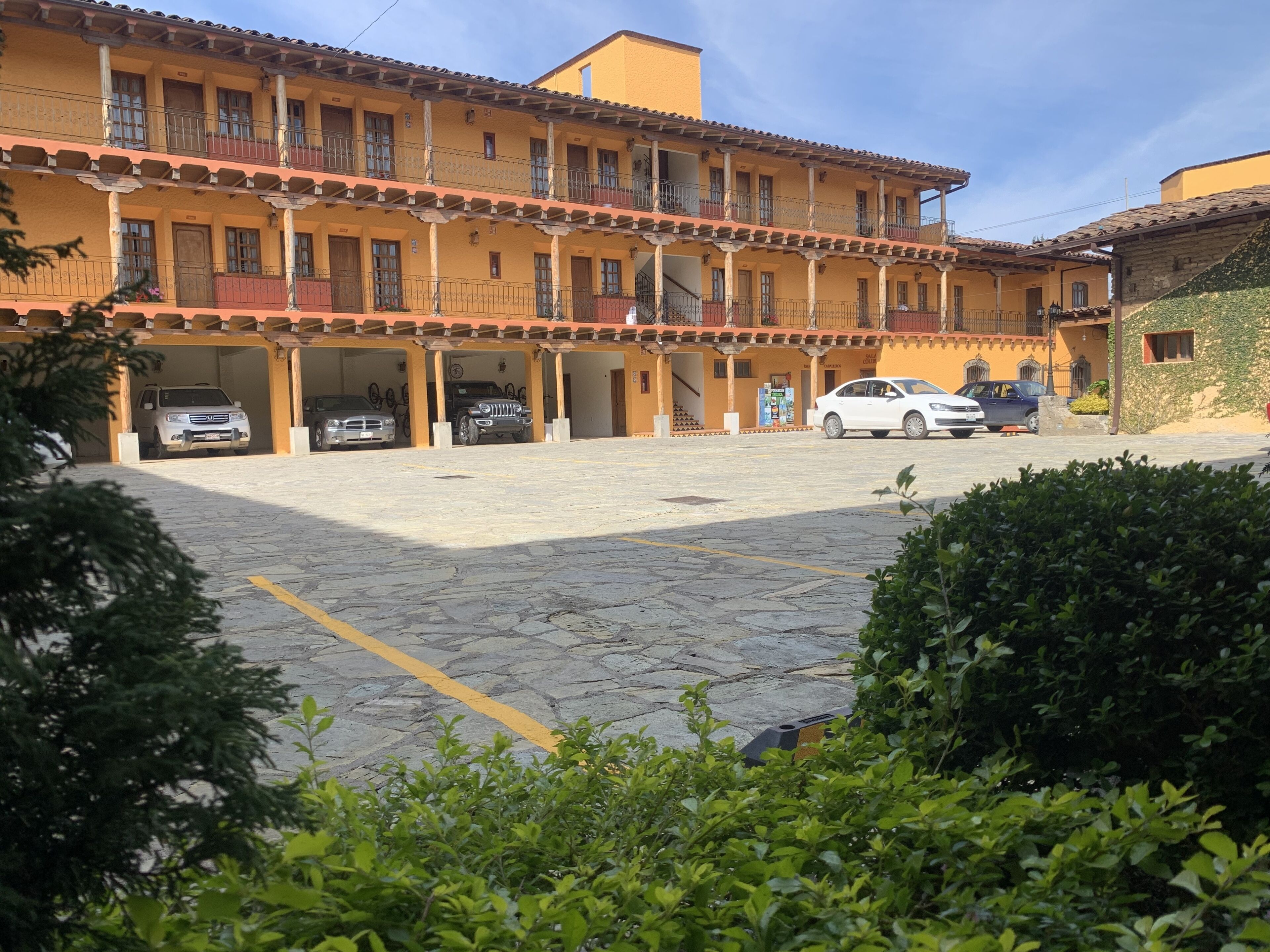 A courtyard with a building featuring a tiled roof and multiple balconies. There are several cars parked in the area, and the ground is paved with stone tiles.