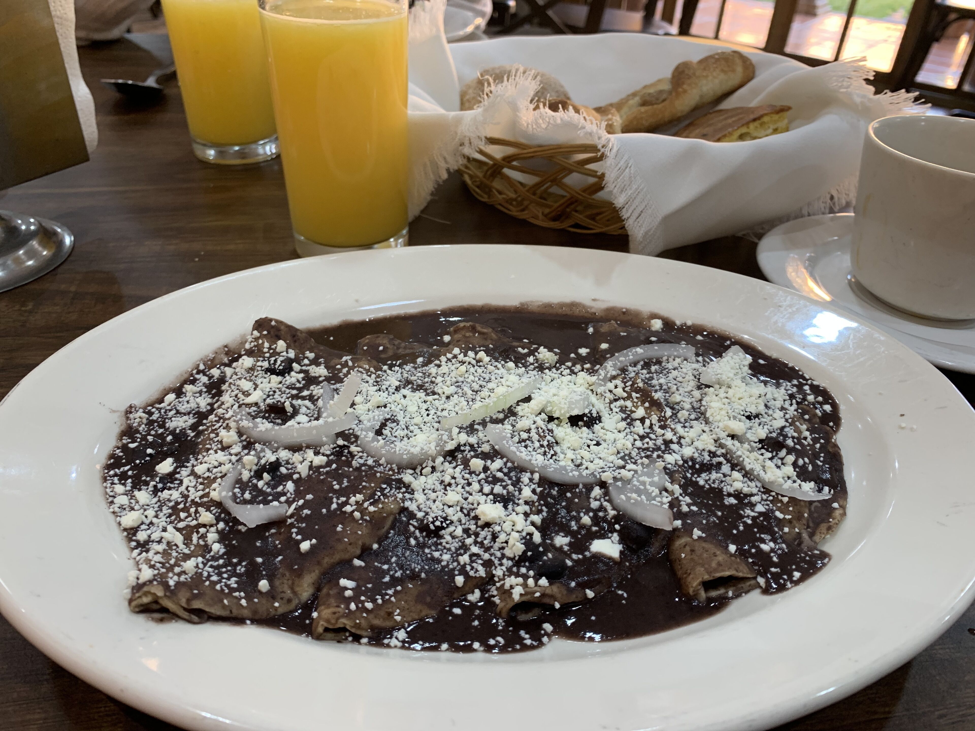 Un plato de crepes de chocolate con acompañamientos, junto a una cesta de pan y un vaso de jugo de naranja.