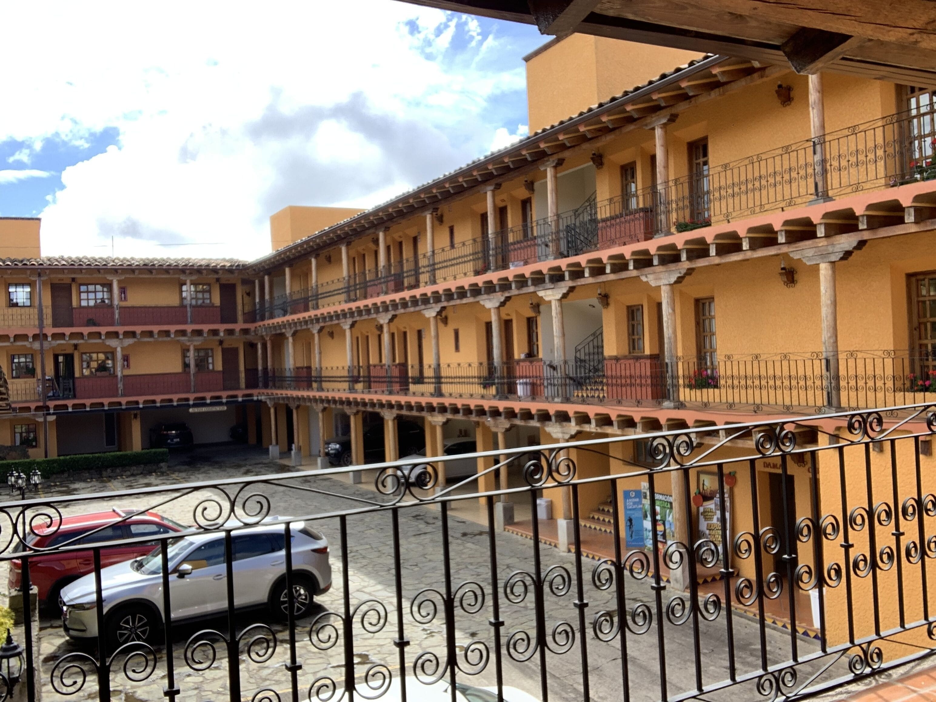 A courtyard with a two-story building, featuring balconies with wrought iron railings, a paved ground, and parked cars.
