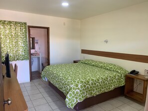 A bedroom with a bed, a wooden nightstand, a desk, and a bathroom visible through an open door.