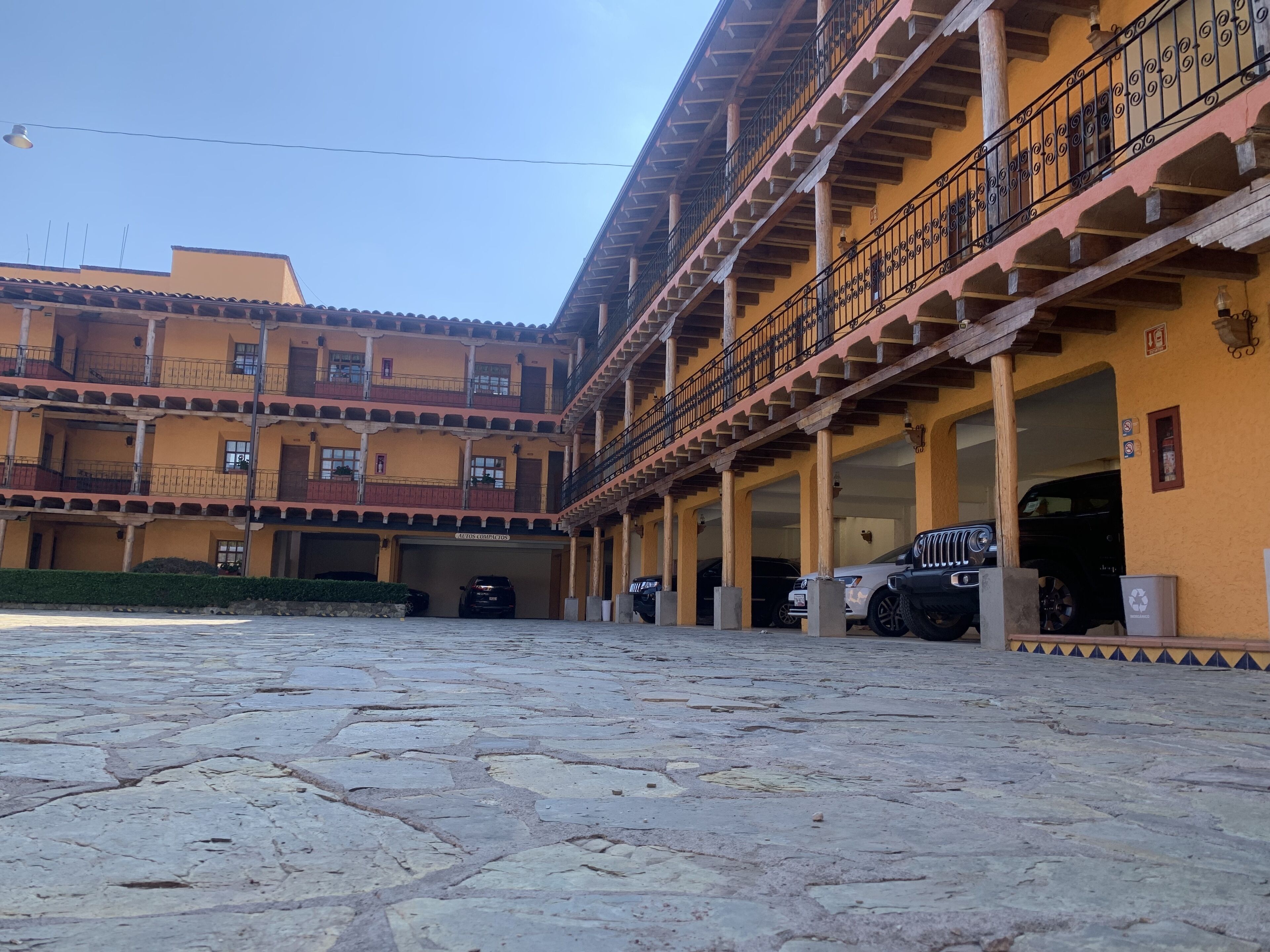 Un patio con piso de piedra, un edificio con varios balcones y una escalera, y varios coches aparcados.